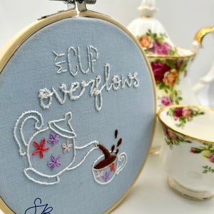 May include: Embroidered hoop art with the words "Cup Overflows" and a teapot pouring into a teacup. The design is on a light blue fabric, with a wooden hoop. A floral teacup and teapot are in the background.