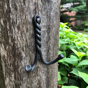 Pode incluir: Um gancho de metal preto com um design retorcido é fixado a um poste de madeira. O gancho tem duas pontas e é feito de ferro forjado.