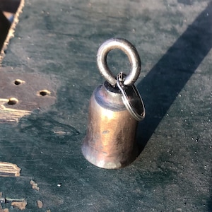 May include: A small, silver bell with a ring attached to the top. The bell is sitting on a green surface.