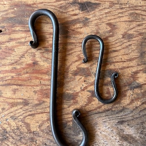Peut inclure: Deux crochets en S en métal foncé forgés à la main de différentes tailles reposent sur une surface en bois. Le crochet le plus grand est allongé, tandis que le plus petit est plus compact. Les deux ont des extrémités recourbées.