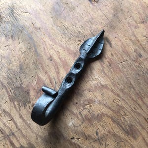 May include: Hand-forged metal leaf-shaped hook. The hook is dark gray with a leaf design at the top and a curved hook at the bottom. Two holes are drilled into the stem of the leaf. The hook is designed to be mounted on a wall or other surface.