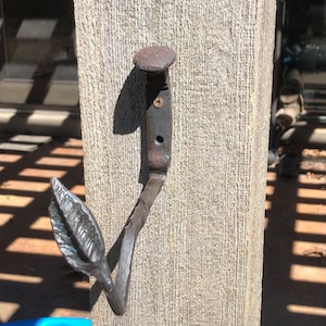 Peut inclure: Un crochet en métal rustique avec un motif de feuille. Le crochet est en métal foncé et a une finition martelée. Il est fixé à une surface en bois.