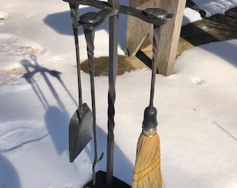 Puede incluir: Un juego de herramientas para chimenea de metal negro con un cepillo, una pala y un atizador. Las herramientas están montadas en un soporte con un diseño de metal retorcido. El juego está sentado sobre una superficie nevada.