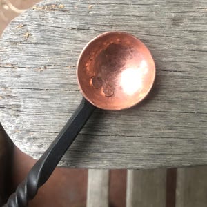 Könnte beinhalten: Ein handgefertigter Kupferlöffel mit einer runden Schüssel und einem gedrehten schwarzen Griff. Die Schüssel des Löffels hat eine polierte, reflektierende Oberfläche. Der Löffel ist für den Küchengebrauch konzipiert.