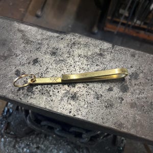 May include: A brass key holder with a loop and a curved bar. The key holder has a small emblem on the bar. The key holder is lying on a textured metal surface.