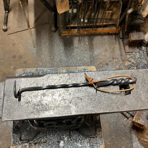 May include: A hand-forged, black metal blacksmith's tool with a twisted handle and a hooked end. A brown leather strap is attached to the handle. The tool rests on a metal anvil.