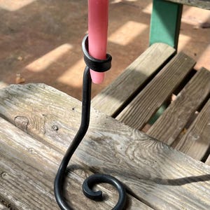 May include: A handcrafted, black metal candle holder with a curled base and a loop to hold a pink candle. The candle is lit, with a yellow flame. The candle holder is on a wooden surface.
