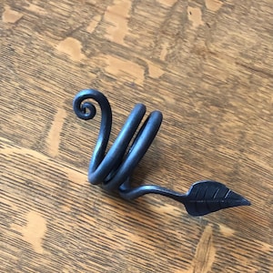 May include: A black metal napkin ring with a leaf design. The ring is coiled and has a leaf shape on the end.