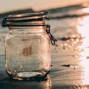 May include: A clear glass jar with a metal lid and a wire handle. The jar is sitting on a sandy beach with the sun setting in the background.
