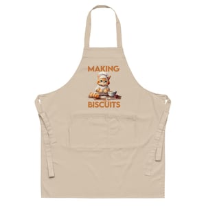 Puede incluir: Un delantal de cocina beige con un bolsillo grande. El delantal tiene un gráfico de un gato con un gorro de chef haciendo galletas. El texto "MAKING BISCUITS" está impreso debajo del gato.
