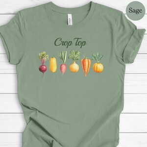 May include: Sage green crop top t-shirt with the words "Crop Top" in a script font. Below the text is a watercolor illustration of various vegetables, including a beet, squash, carrot, onion, and pepper.