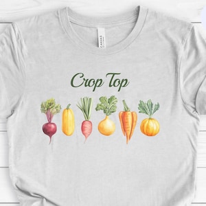 An ash grey crewneck shirt laying flat against a white wooden backdrop. The design is located at the center chest and features the food pun phrase "Crop Top" with a row of colorful, vintage style vegetable illustrations underneath.