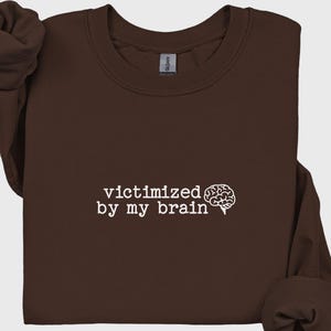 May include: A dark brown sweatshirt with the phrase "victimized by my brain" printed in white, accompanied by a white brain graphic. The sweatshirt has a crew neck and long sleeves.