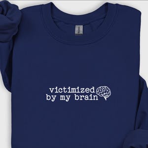 May include: A navy blue sweatshirt with the phrase "victimized by my brain" in white text, accompanied by a brain graphic. The sweatshirt is made of a soft material and has a crew neck.