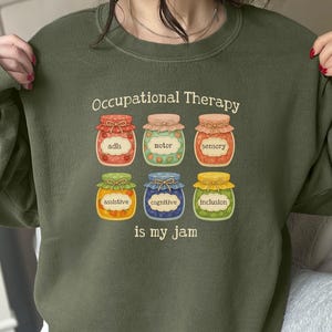 May include: Olive green sweatshirt with the text "Occupational Therapy is my jam." The design features six jars with labels like "ADLs," "Motor," and "Sensory." The jars are in red, green, pink, orange, blue, and yellow.