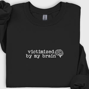 May include: Black sweatshirt with the text "victimized by my brain" in white, accompanied by a white brain graphic. The sweatshirt is made of a soft material and has a crew neck.
