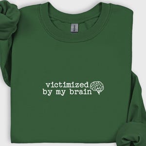 May include: A dark green sweatshirt with the phrase "victimized by my brain" in white text, accompanied by a white brain graphic. The sweatshirt has long sleeves and a crew neck.