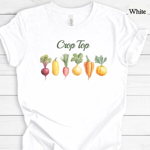 May include: White crop top t-shirt with a watercolor illustration of various vegetables, including a beet, zucchini, carrot, turnip, pepper, and pumpkin. The words "Crop Top" are printed in green cursive above the vegetables.