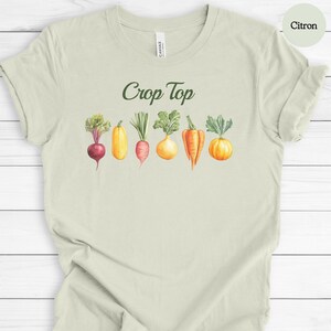 May include: Citron-colored t-shirt with the words "Crop Top" in green script. Below the text is a watercolor illustration of various vegetables, including a beet, squash, carrot, and pumpkin. The shirt is made of a soft material.