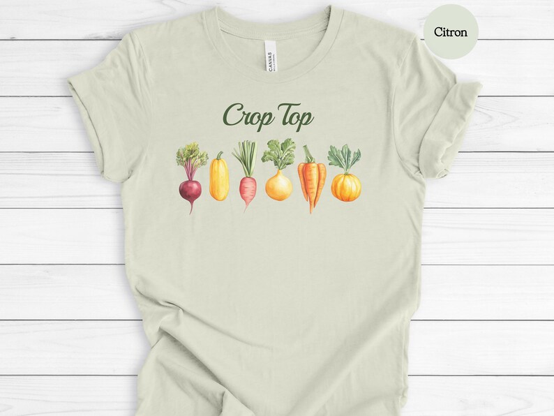 May include: Citron-colored t-shirt with the words "Crop Top" in green script. Below the text is a watercolor illustration of various vegetables, including a beet, squash, carrot, and pumpkin. The shirt is made of a soft material.