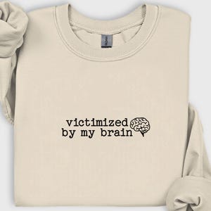 May include: A cream-colored sweatshirt with the phrase "victimized by my brain" printed in black, accompanied by a brain graphic. The sweatshirt has a crew neck and long sleeves, folded neatly. The Gildan brand tag is visible.