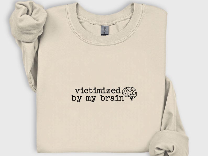 May include: A cream-colored sweatshirt with the phrase "victimized by my brain" printed in black, accompanied by a brain graphic. The sweatshirt has a crew neck and long sleeves, folded neatly. The Gildan brand tag is visible.