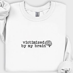 May include: White crewneck sweatshirt with the phrase "victimized by my brain" in black text, accompanied by a brain graphic. The sweatshirt has long sleeves and a casual style.