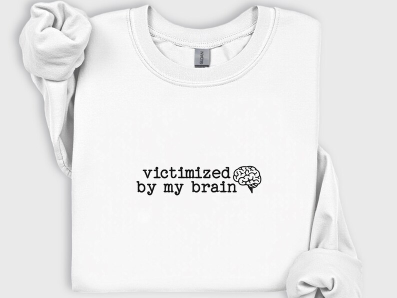 May include: White crewneck sweatshirt with the phrase "victimized by my brain" in black text, accompanied by a brain graphic. The sweatshirt has long sleeves and a casual style.
