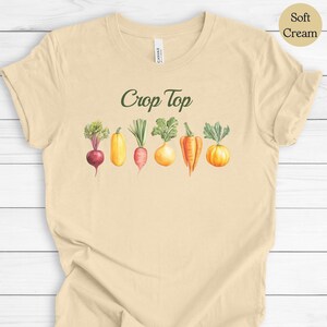 May include: A soft cream-colored crop top featuring the text "Crop Top" in green script above a watercolor illustration of various vegetables, including a beet, squash, carrot, and onion.