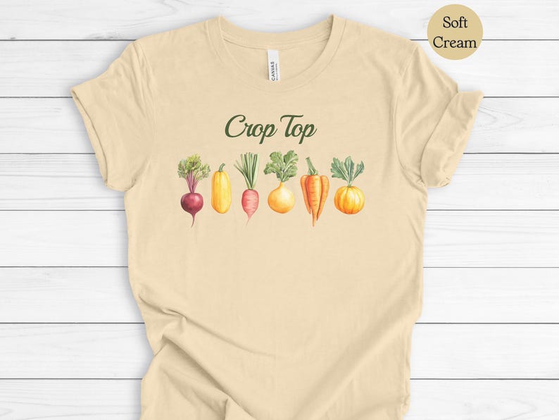 May include: A soft cream-colored crop top featuring the text "Crop Top" in green script above a watercolor illustration of various vegetables, including a beet, squash, carrot, and onion.