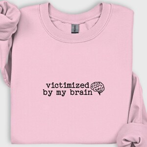 May include: A light pink sweatshirt with the phrase "victimized by my brain" printed in black, along with a brain graphic. The sweatshirt has a crew neck and long sleeves, with the sleeves rolled up.