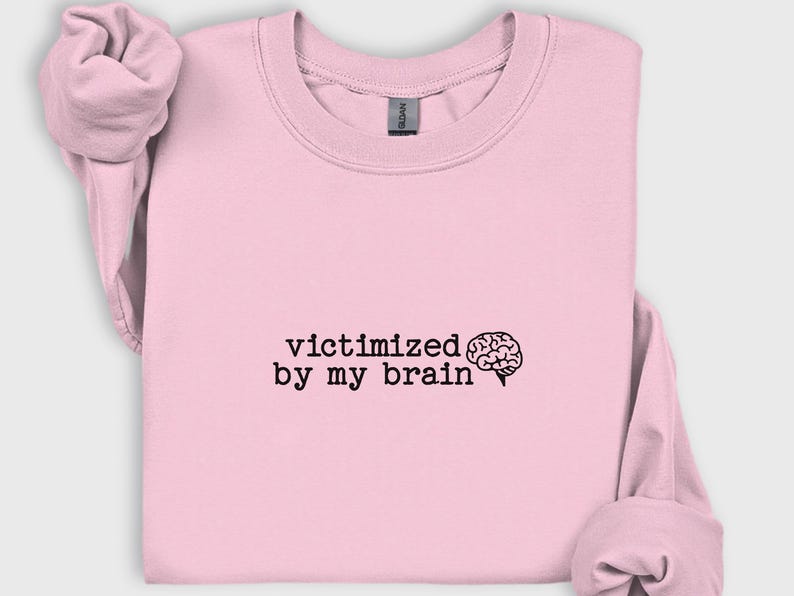 May include: A light pink sweatshirt with the phrase "victimized by my brain" printed in black, along with a brain graphic. The sweatshirt has a crew neck and long sleeves, with the sleeves rolled up.