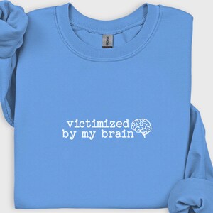 May include: A light blue sweatshirt with the phrase "victimized by my brain" printed in white, along with a white brain graphic. The sweatshirt has long sleeves and a crew neck.