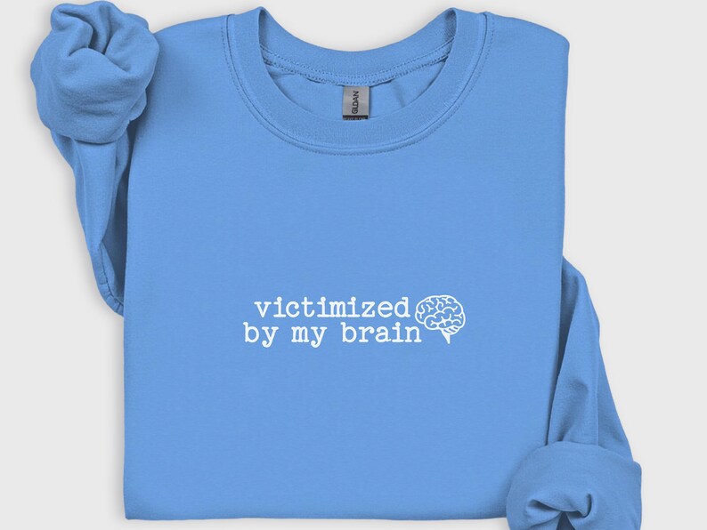 May include: A light blue sweatshirt with the phrase "victimized by my brain" printed in white, along with a white brain graphic. The sweatshirt has long sleeves and a crew neck.