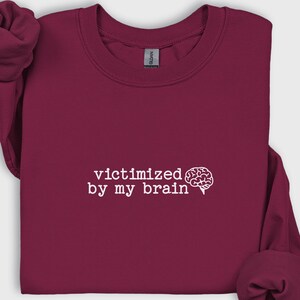 May include: A maroon sweatshirt with the phrase "victimized by my brain" in white text, accompanied by a white brain graphic. The sweatshirt has long sleeves and a crew neck.