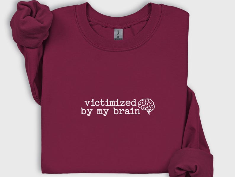 May include: A maroon sweatshirt with the phrase "victimized by my brain" in white text, accompanied by a white brain graphic. The sweatshirt has long sleeves and a crew neck.
