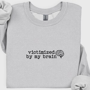 May include: Light gray sweatshirt with the phrase "victimized by my brain" in black text, accompanied by a brain graphic. The sweatshirt has a crew neck and long sleeves.