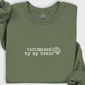 May include: Olive green sweatshirt with the phrase "victimized by my brain" in white, accompanied by a brain graphic. The long-sleeved top is made of a soft material.