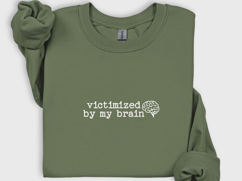 May include: Olive green sweatshirt with the phrase "victimized by my brain" in white, accompanied by a brain graphic. The long-sleeved top is made of a soft material.