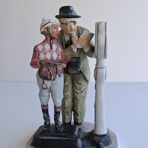 May include: A ceramic figurine depicting a jockey in a pink and white diamond pattern shirt and white pants, and an official in a suit and hat, standing near a scale. The figurine is on a rectangular base.