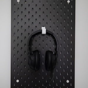 May include: A black pegboard with a grid of vertical slots is mounted on a wall. A white plastic hook is inserted into the centre of the board, holding a pair of black over-ear headphones with a microphone boom attached to the right side.