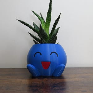 Op de afbeelding: Een blauwe, lachende, karaktervormige plantenbak met een rode mond en zwarte ogen, met een groene vetplant. De plantenbak staat op een houten oppervlak.