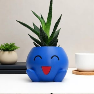 Peut inclure: Un pot de fleurs sphérique bleu vif arborant un visage de dessin animé avec deux yeux noirs incurvés et une bouche triangulaire rouge. Le pot possède deux petits pieds arrondis à la base et contient une plante succulente verte épineuse.
