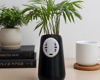 May include: A black cylindrical planter featuring a white mask-like face with purple markings. The planter holds a green palm plant. The base of the planter is rounded, and the surface is smooth. The design resembles a character from Spirited Away.