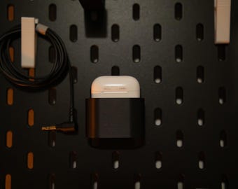 May include: A black pegboard with a white wireless earbud case in a black holder. A black cable and a white clip are on the left side. Another white clip is on the right side. The pegboard has oval-shaped holes.