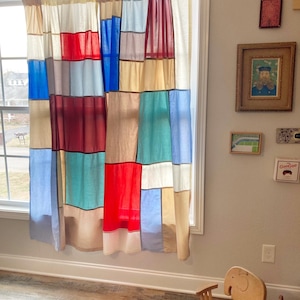 May include: A pair of patchwork curtains featuring a grid of rectangular fabric panels in various colors including red, blue, teal, cream, and tan. The curtains are hung on a black rod over a window, displaying a multi-colored block pattern.