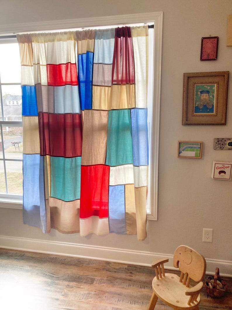 May include: A pair of patchwork curtains featuring a grid of rectangular fabric panels in various colors including red, blue, teal, cream, and tan. The curtains are hung on a black rod over a window, displaying a multi-colored block pattern.