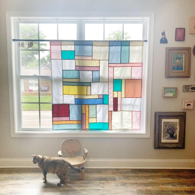 May include: A rectangular window curtain featuring a grid of multicolored fabric panels in shades of teal, yellow, pink, blue, and white. The curtain is suspended from a dark rod and displays a geometric patchwork design with varied rectangular shapes.