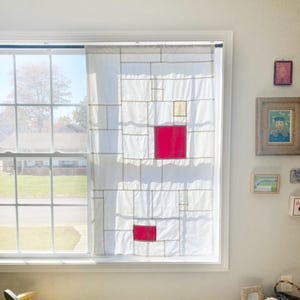 May include: A white window curtain featuring a geometric grid pattern composed of thin tan lines, with two prominent solid red squares and one smaller solid beige square integrated into the design, hanging from a dark rod over a window pane.