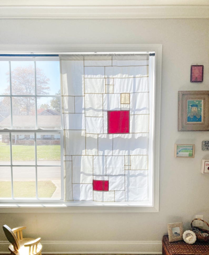 May include: A white window curtain featuring a geometric grid pattern composed of thin tan lines, with two prominent solid red squares and one smaller solid beige square integrated into the design, hanging from a dark rod over a window pane.